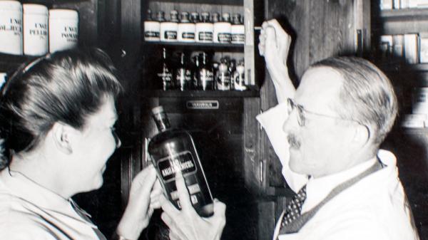 Eine Frau steht gemeinsam mit Wilhelm Seyd vor einem historischen Apothekerschrank. Sie schauen auf eine Flasche mit Arznei, die der Mann in der Hand hält. Das Bild ist schwarz-weiß.