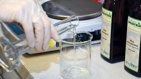 Eine Hand mit Gummihandschuh gießt eine Flüssigkeit aus einem Reagenzglas in ein gläsernes Gefäß. Daneben stehen eine Waage und zwei Flaschen mit der Aufschrift „Spagyrik“.