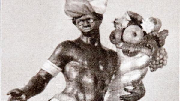 Schwarz-weiß-Bild einer männlichen Figur mit dunkler Hautfarbe und nacktem Oberkörper. Sie trägt einen Turban und hält ein Horn mit Obst im Arm. Die Darstellung widerspiegelt die Sichtweise der europäischen Länder auf dunkelhäutige Menschen zur Zeit des Kolonialismus.